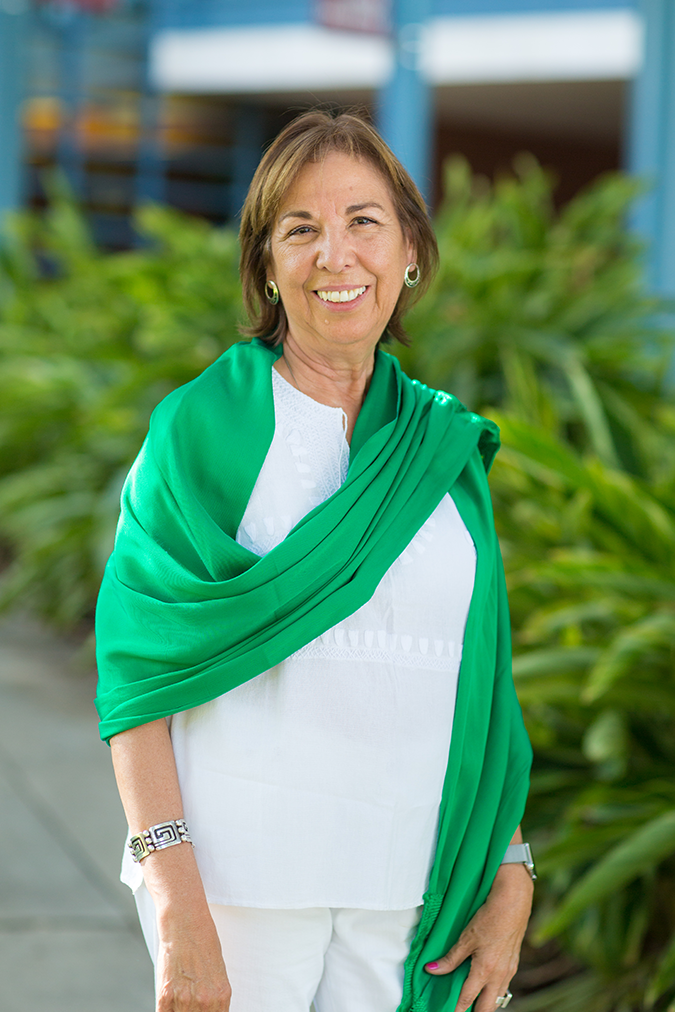 A woman stands, smiling at the camera, dressed in white and draped in a bright green scarf.