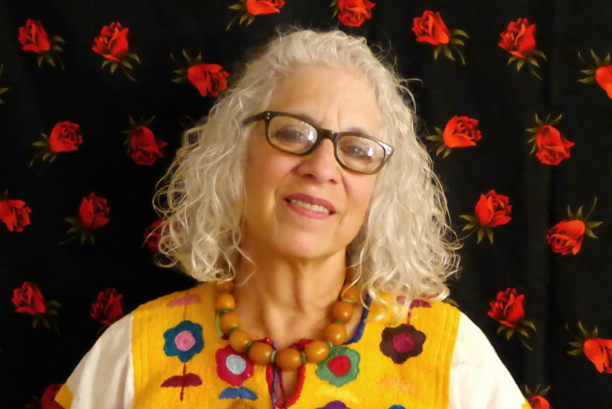 A headshot of a woman in eyeglasses, wearing a bright yellow top with a flower pattern, smiling at the camera.