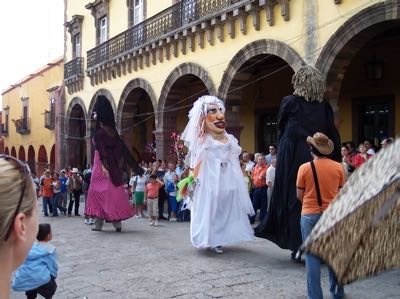 Giant masked figures in costume, including a female figure in a white veil and dress, in a courtyard with a crowd watching from the periphery