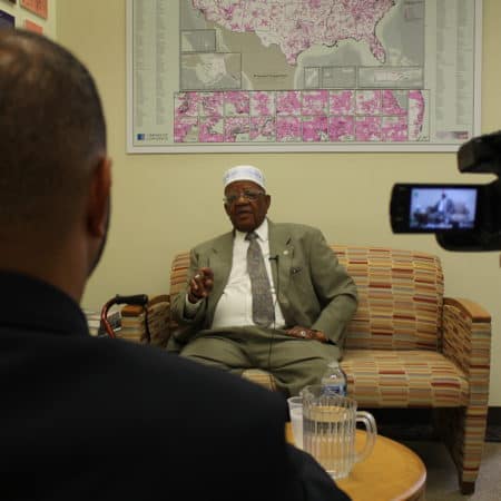 We see over the shoulder of an interviewer and a videorecorder pointed at a man seated on a couch