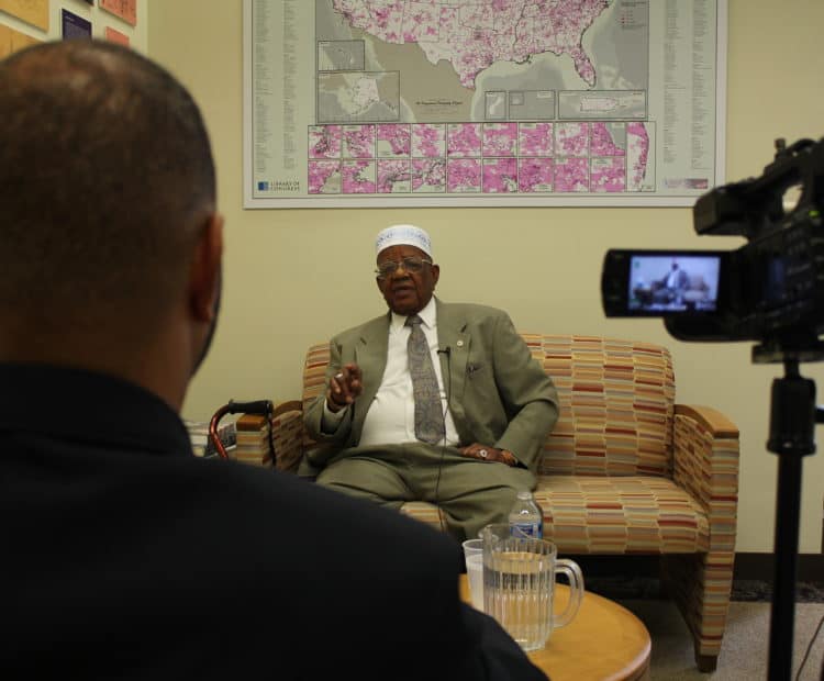 We see over the shoulder of an interviewer and a videorecorder pointed at a man seated on a couch