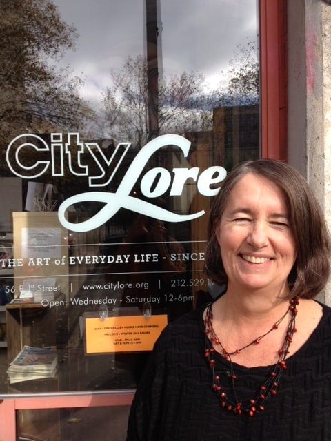 A headshot of a woman, standing in front of a storefront window, smiling at the camera.