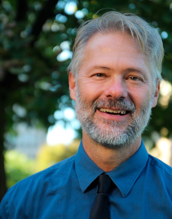 A headshot of a man, wearing a royal blue shirt with a navy blue tie, smiles at the camera.