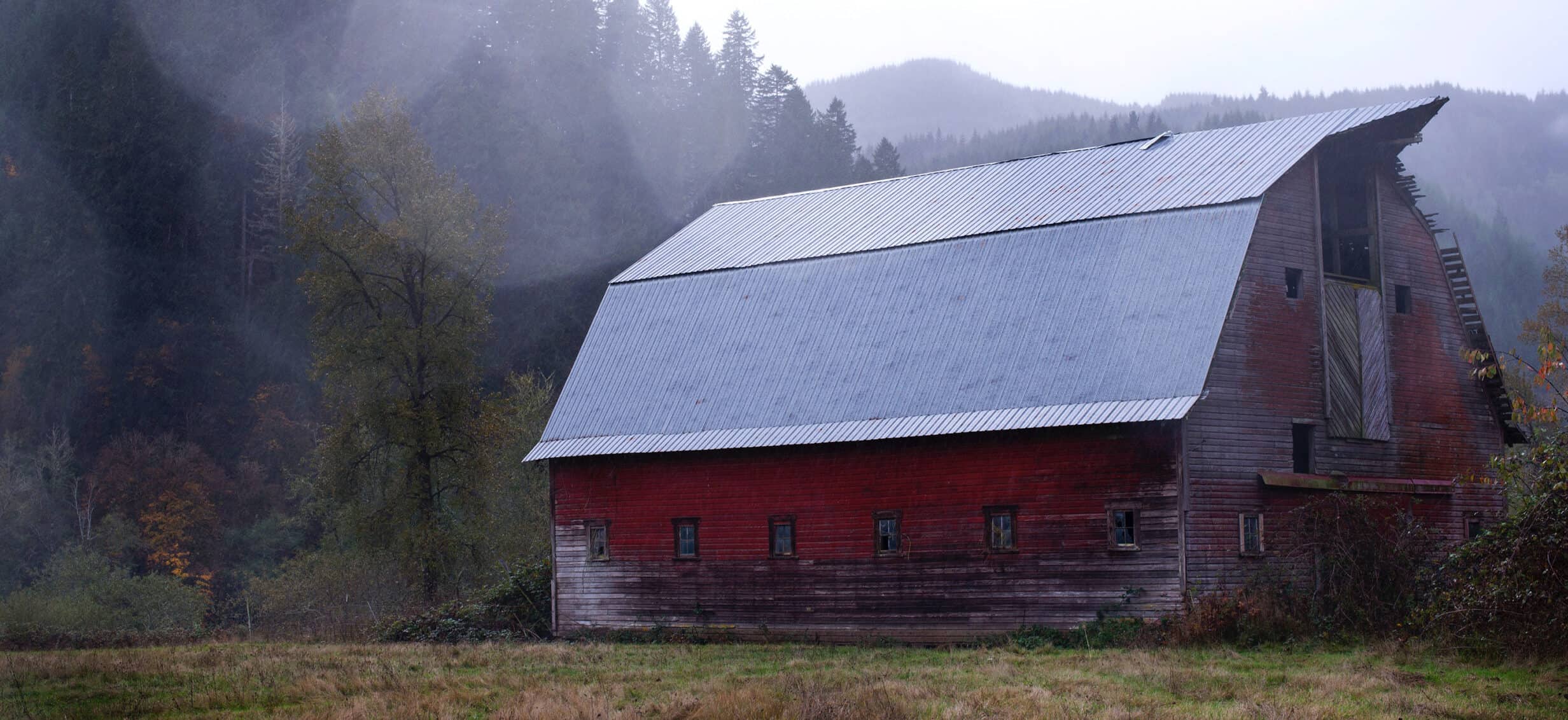 A large, red barn with a small mountain in the background and fog in the air.