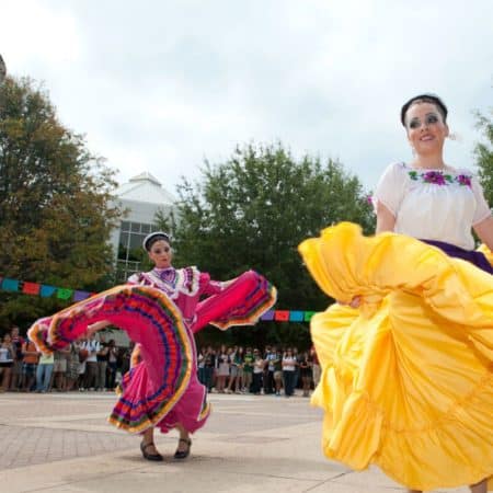 A man in a black embroidered suit dances next to three women in colorful dresses.