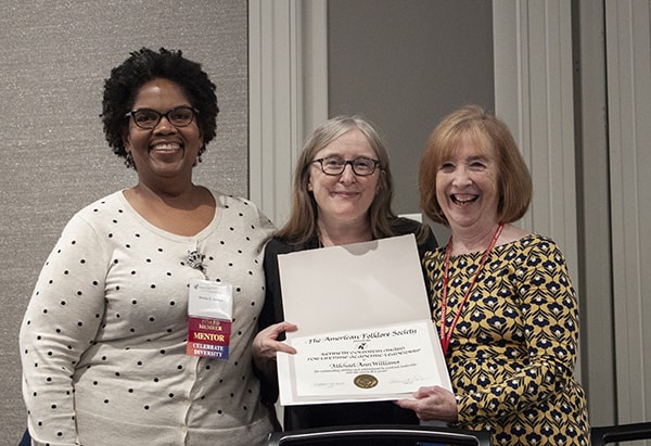 Three women stand, smiling at the camera, the center woman holds an award certificate.