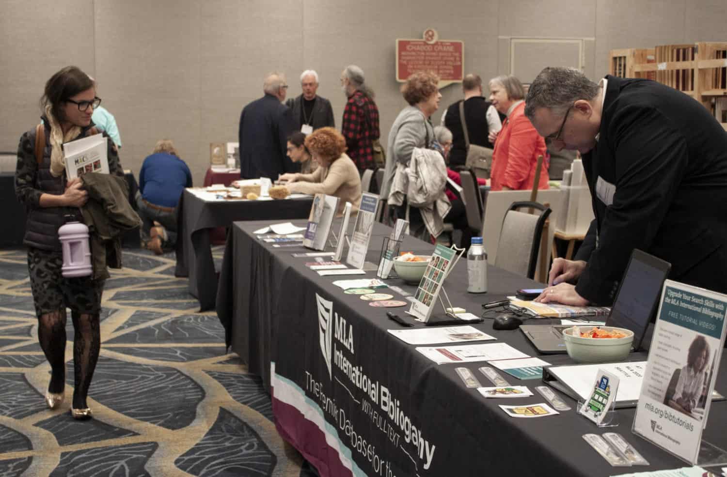 People browse and chat in groups around display tables in a conference exhibit room.