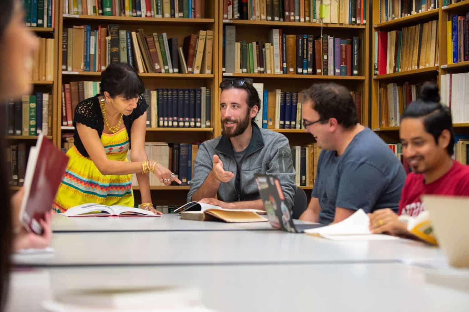A female teacher, standing, instructs three men sitting at a table.