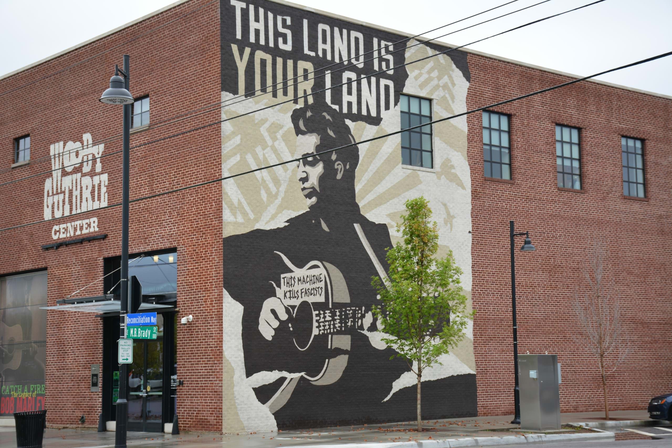 A mural of folk musician Woody Guthrie featuring an image of Guthrie playing guitar, below the text "The Land is Your Land".