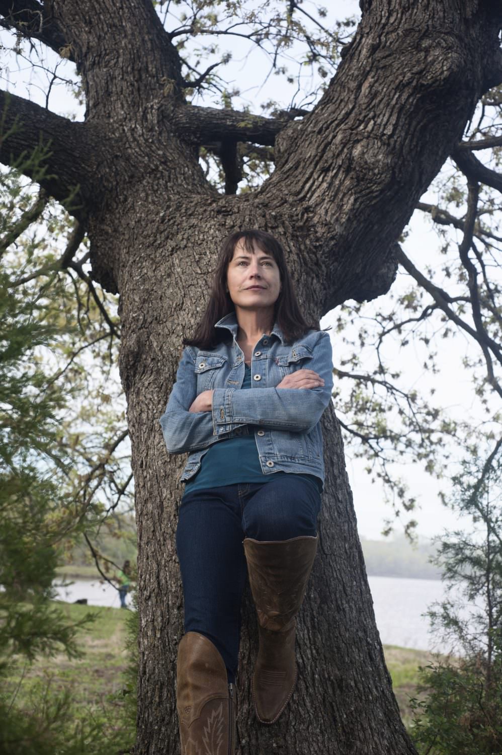 Photo of Mish leaning against a large tree. Mish's left leg is propped against the tree, and is wearing a jean jacket and dark blue jeans.