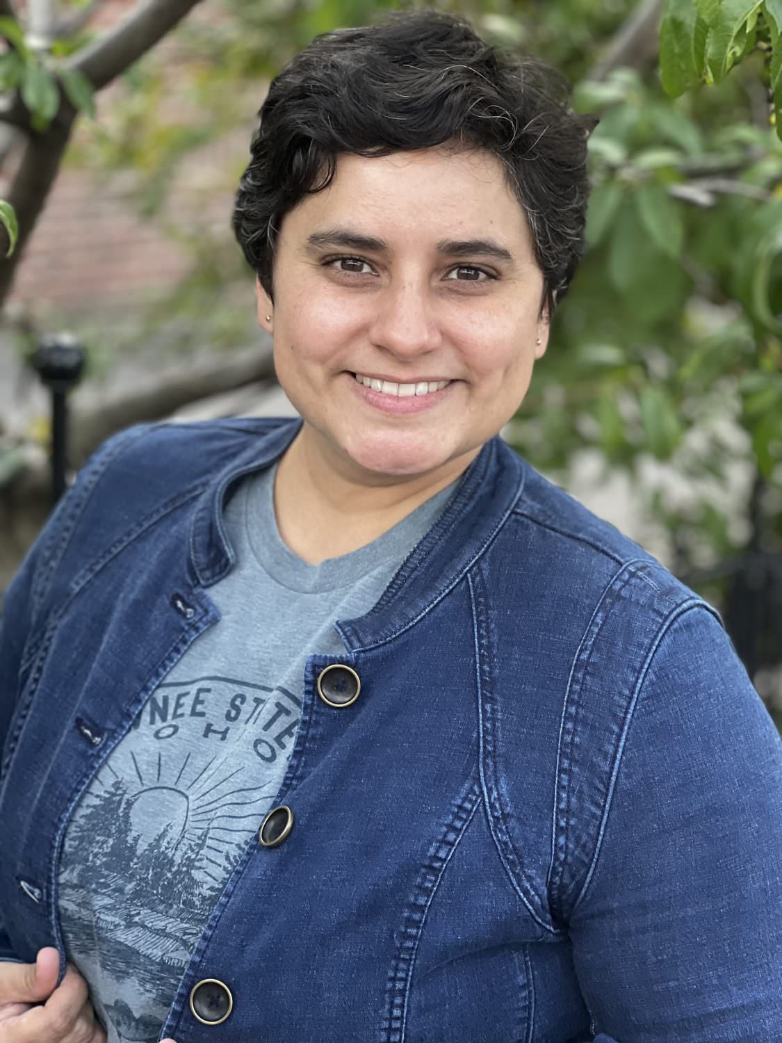 ID: Headshot of Dr. Cassie Rosita Patterson, who has olive-colored skin, short and wavy dark brown hair (with some whites coming in along the front), and is wearing a blue shirt and blue denim jacket. 