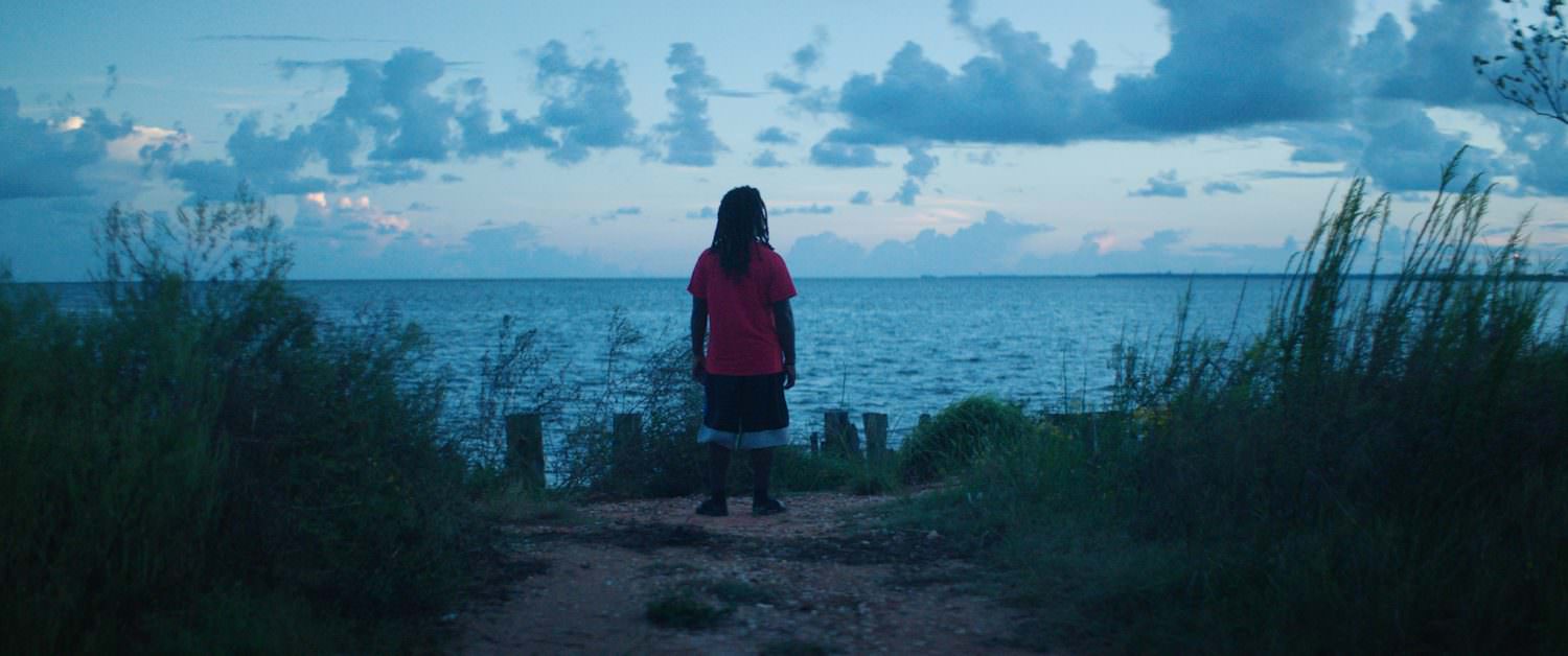Emmett Lewis standing at a dock overlooking a body of water and horizon. Lighting is blue and green, and Lewis is wearing a red shirt and black shorts with a white hem.