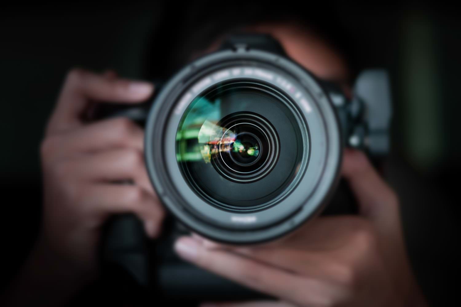 A camera lens facing viewer, with hands holding camera in the background