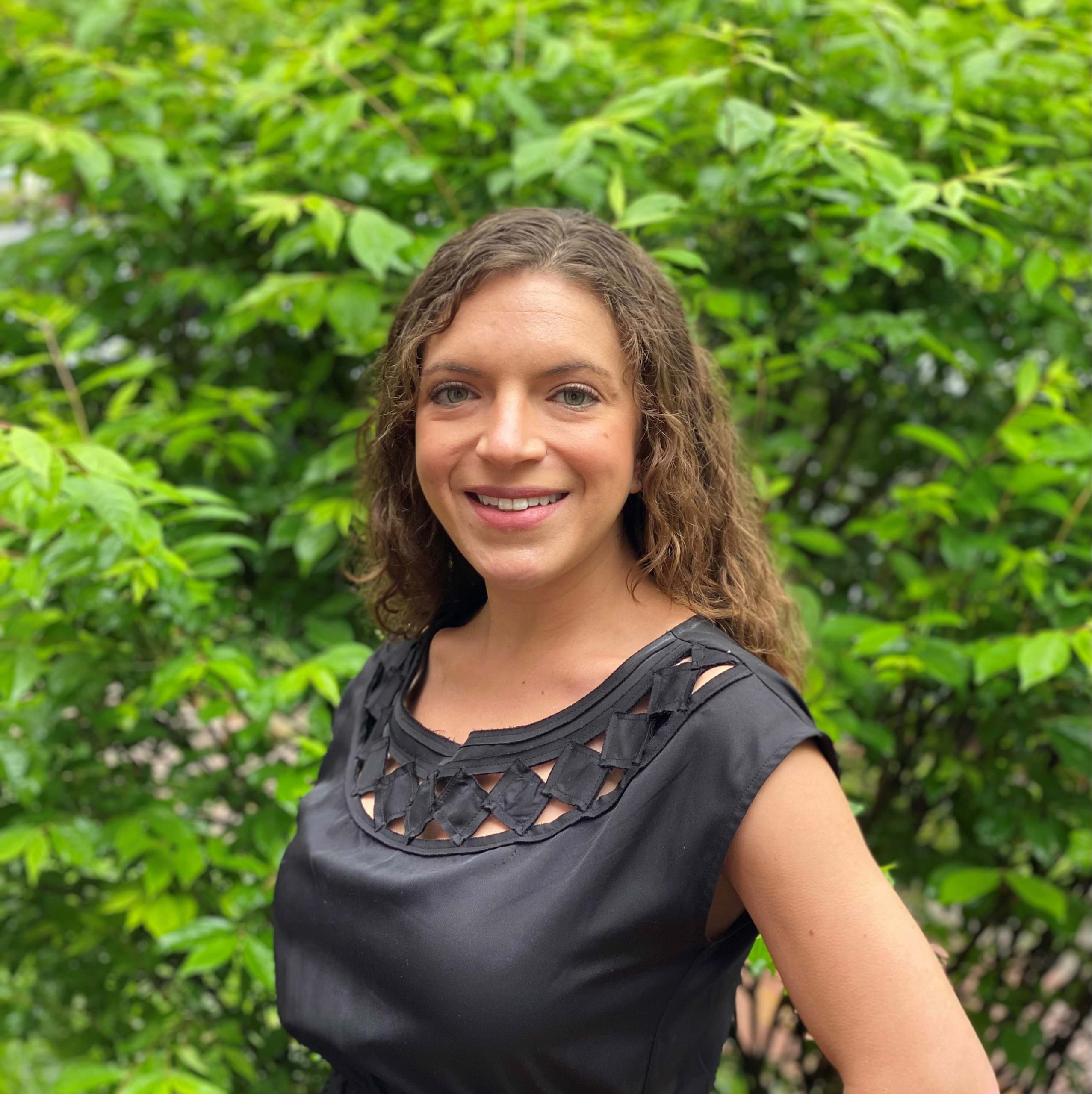 headshot of Rachel McKean, who wears a dark grey shirt with a geometric cutout pattern around the neck and stands in front of greenery
