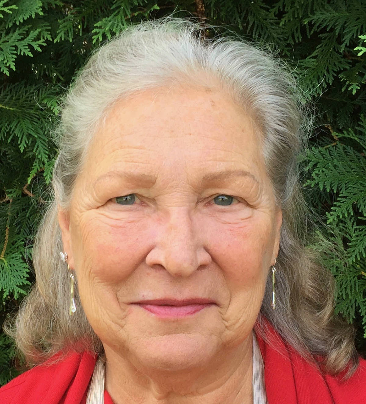 The photo is a headshot of Bonnie Blair O'Connor, taken against a backdrop of evergreen fronds. In this photo Dr. O'Connor wears a bright red jacket and silver earrings and has shoulder-length, mostly grey-white hair.