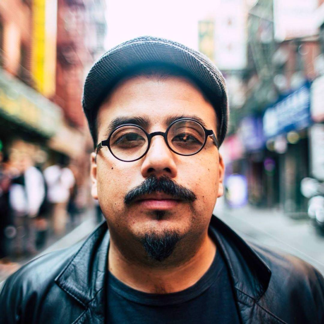 bayona on a city street wearing a leather jacket, glasses, and cap