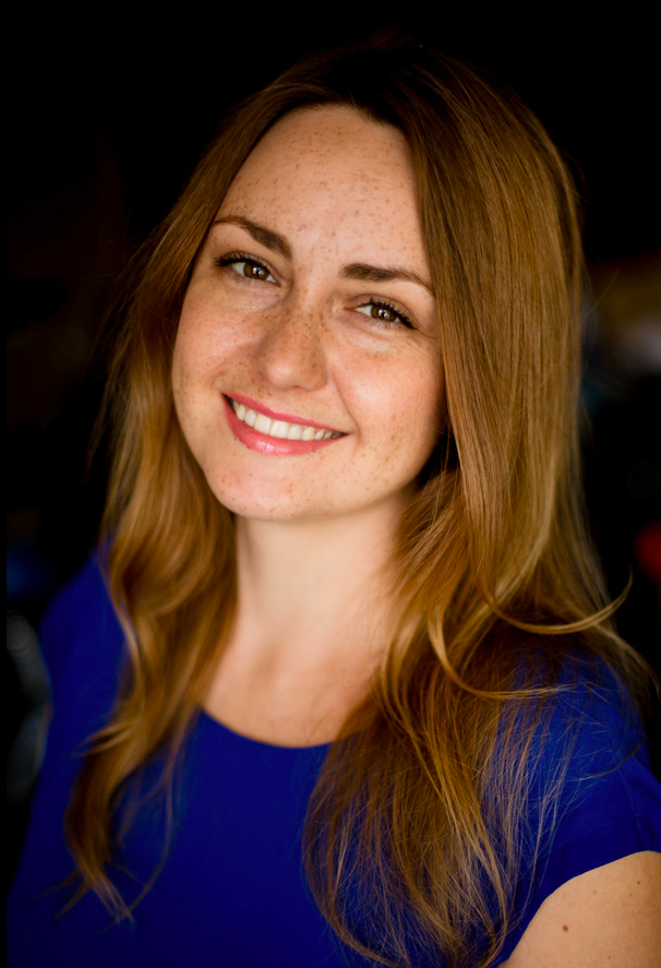 Iryna has light brown long wavy hair and brown eyes. She is wearing a dark blue blouse. 