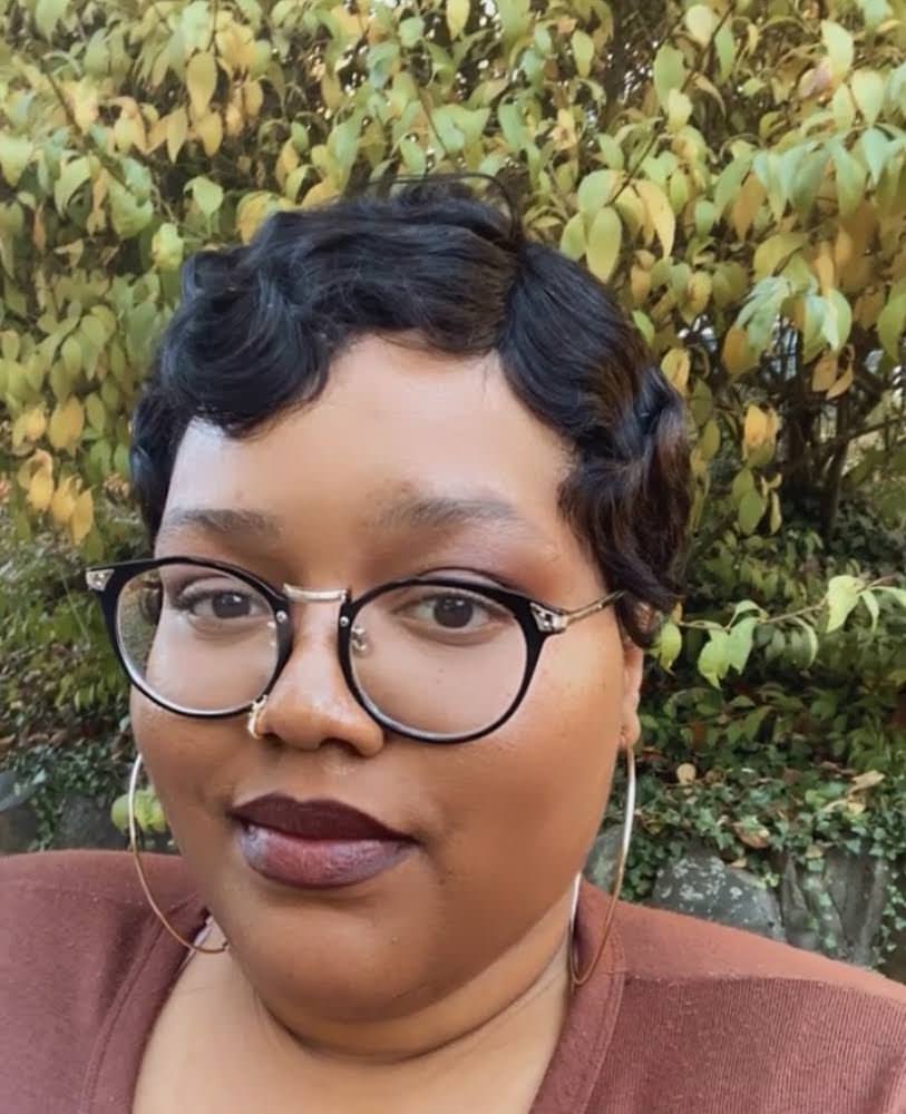 light brown skin Black person wearing glasses, a brown shirt, with short dark hair in finger waves in front of a fall foliage.