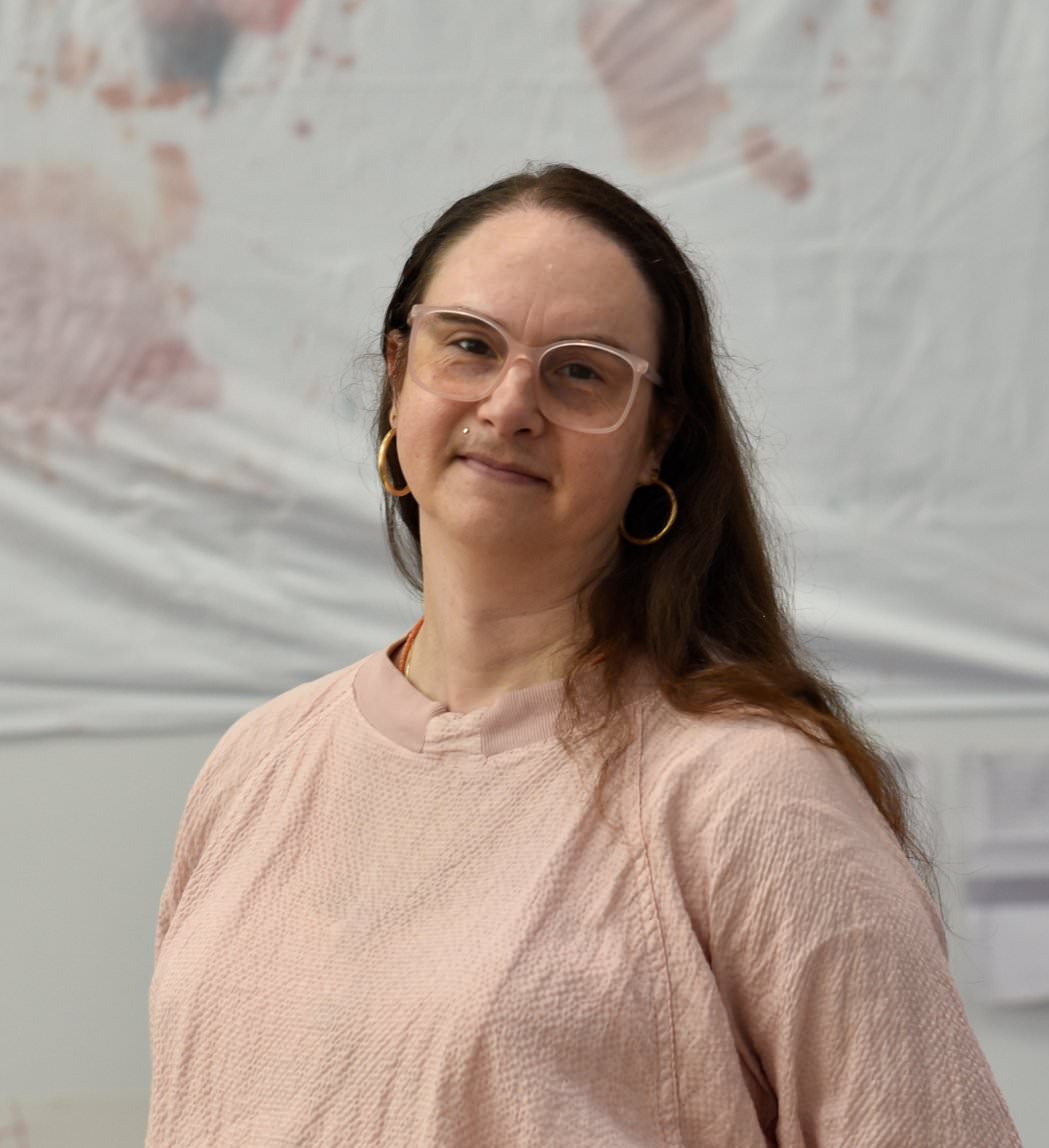 A photograph of Gavilán Rayna Russom by Elodie Adam. Rayna appears from mid chest up and tilts her head to her left as she smiles at the camera. She is a pale skinned woman with long reddish-brown hair parted on the side and a faint mustache. She wears a light pink pullover and large light pink glasses. There is a small gold "monroe" piercing above her right upper lip. She has on gold hoop earrings and a small orange necklace is partially visible poking out of the pullover's collar. She is standing in the main gallery of the Center for Art, Research and Alliances in New York City where she curated an artists residency and exhibition in the summer of 2023. Behind her, slightly out of focus and mounted on a white wall, are works by Omari Love, one of the artists in the exhibition. These include a portion of a white sheet stained with fake blood and ink across the top of the frame and two U.S. letter size sheets of paper with writing on them at the bottom right of the frame.