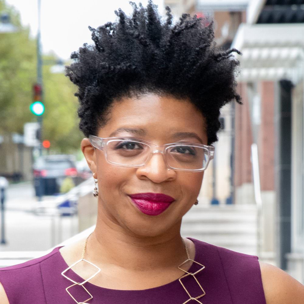 Headshot of Cheryl Schiele, a black woman with short black naturally-kinky hair, is wearing glasses, a purple top, and a gold necklace.