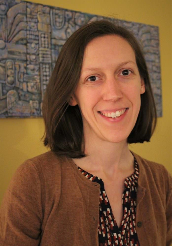 Headshot of Jennie Terman, Folk & Traditional Arts Specialist at the National Endowment for the Arts, who is a white woman with brown hair and brown eyes, wearing a patterned blouse with a brown cardigan and standing in front of a canvas painted by her father.