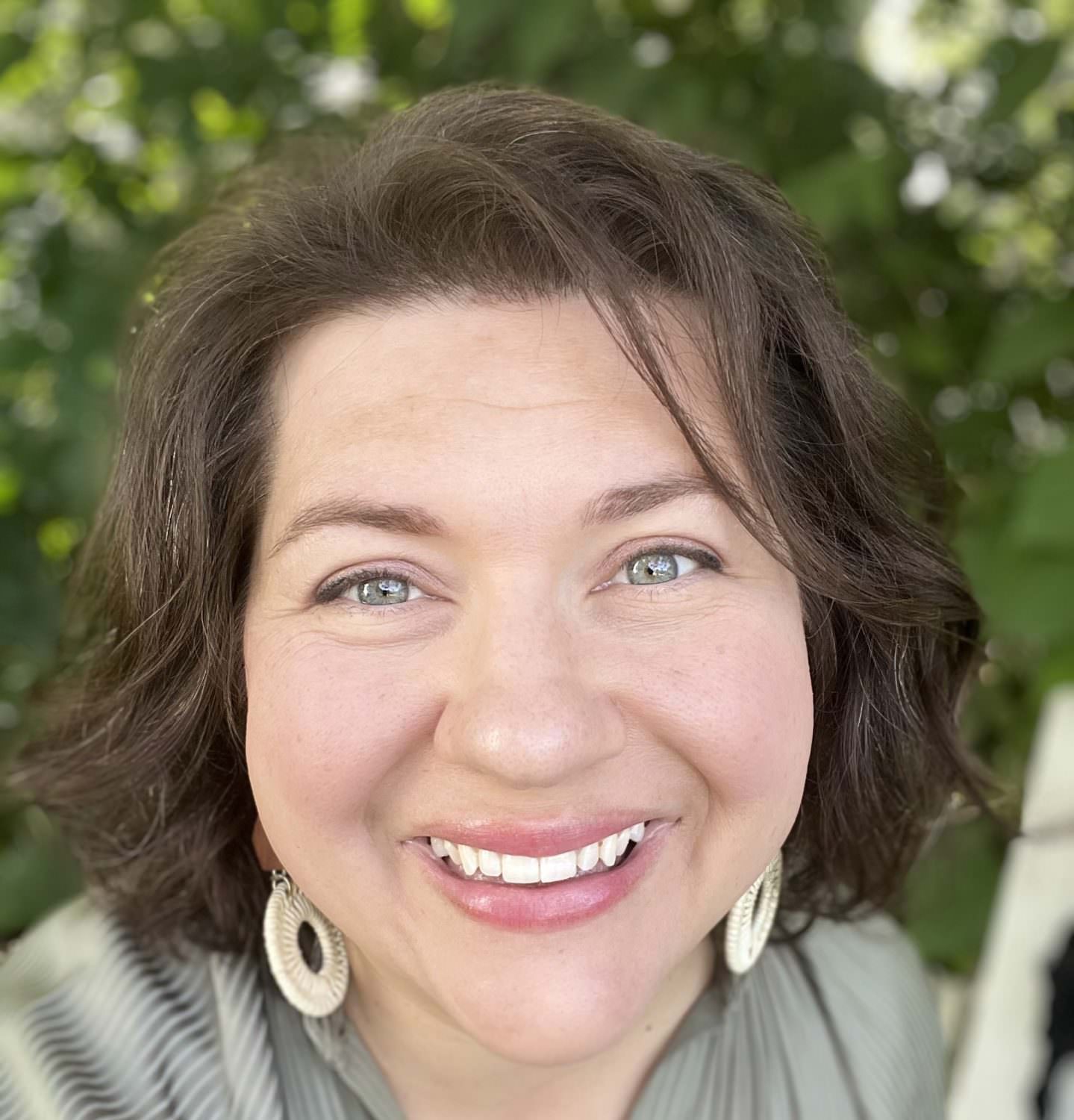 Headshot of caucasian female with dark brown hair, wearing a green blouse while smiling at the camera outside by trees and a white fence.