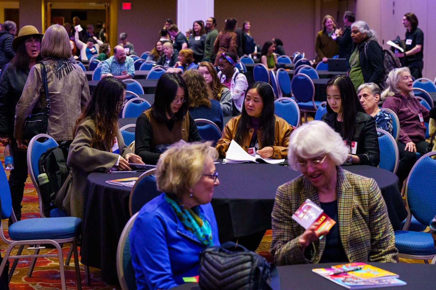 Photo showing round tables and blue chairs in a large room, with more than 20 people sitting or standing there, mostly talking to each other.