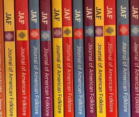A row of JAF issues on a shelf showing the afS logo and the text JAF/Journal of American Folklore. The color of the covers alternate from bright yellow to red, blue and purple before repeating