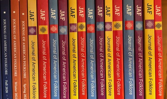 A row of JAF issues on a shelf showing the afS logo and the text JAF/Journal of American Folklore. The color of the covers alternate from bright yellow to red, blue and purple before repeating