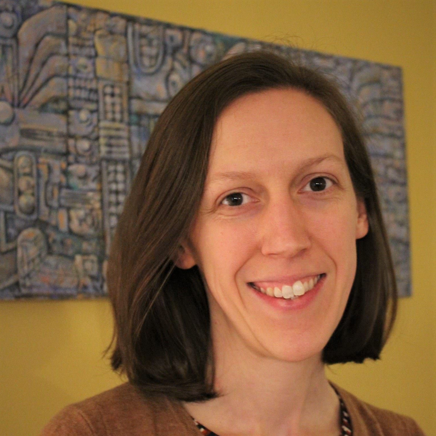 Headshot of Jennie Terman, who is a white woman with chin-length brown hair and brown eyes, wearing a patterned blouse with a brown cardigan and standing in front of a canvas painted by her father.