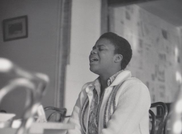 Black woman with short hair and in sweater sitting and singing in a room