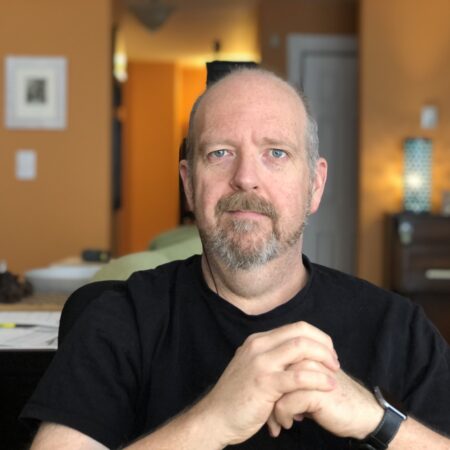 photo of a white male in black shirt and grey beard inside a room saluting the viewers