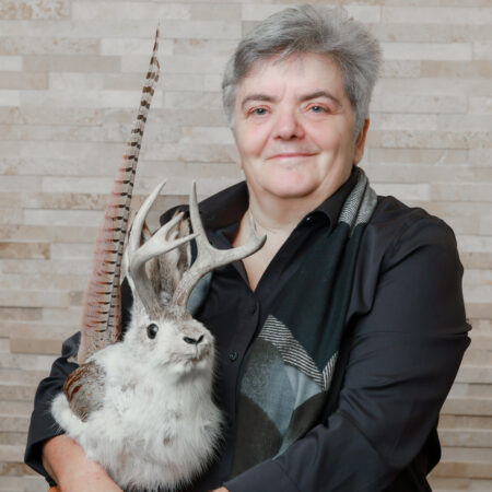headshot of a female in short grey hair holding a taxidermized rodent with antlers and bird feathers