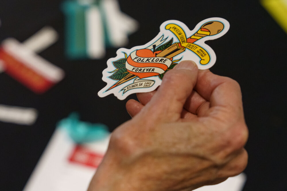 an older person's hand holding the commemorative sticker, which features a peach-colored peach with the words "folklore forever" in a sailor jerry-style dagger and heart tattoo