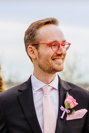 Headshot of Zack Bekowies, Assistant Grants Management Specialist at the National
Endowment for the Arts, who is a white man with short, light-brown hair, green eyes,
and pink glasses wearing a black suit, white collared shirt, light-pink tie, and a pink rose
boutonniere.