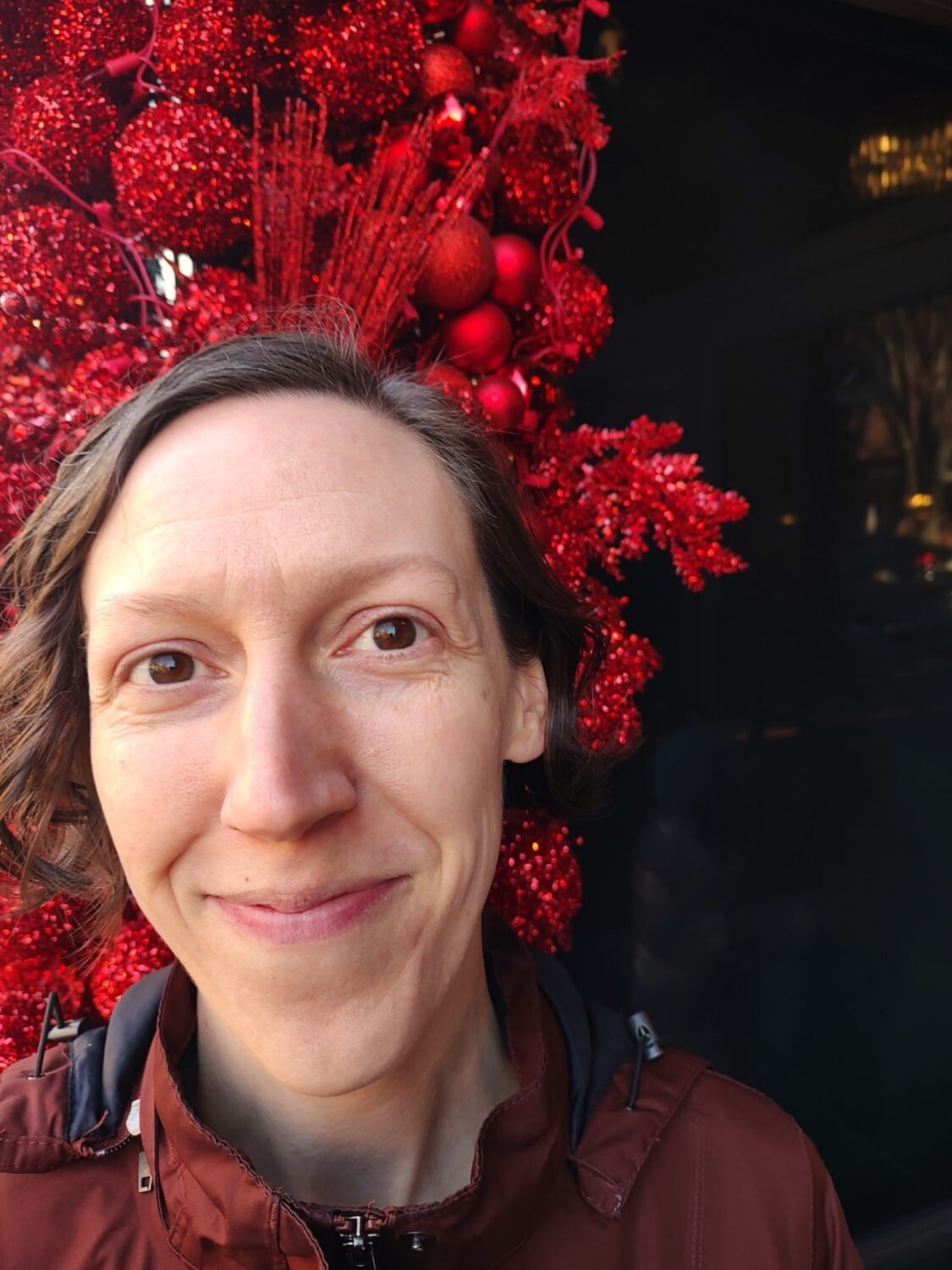 Headshot of Jennie Terman, Folk & Traditional Arts Specialist at the National Endowment for the Arts, who is a white woman with chin-length wavy brown hair and brown eyes, wearing a burgundy-colored jacket and standing in front of red glittery decorations.