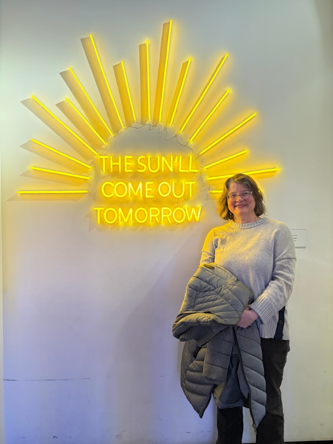 Headshot of Erin Waylor, Coordinator at the National Endowment for the Arts, is a white woman with medium-length brown and grey hair wearing glasses, a grey sweater, and brown pants. She is holding a heavy grey coat in front of a large yellow neon sign that says THE SUN’LL COME OUT TOMORROW.