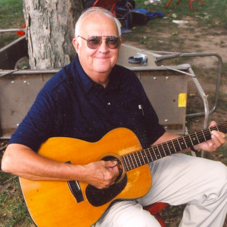 A sitting and smiling white-haired man wearing sun glasses and holding a fiddle